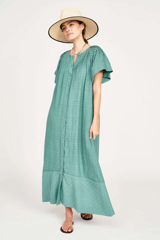 Woman in a flowing teal maxi dress with flutter sleeves, wearing a wide-brimmed hat and sandals.